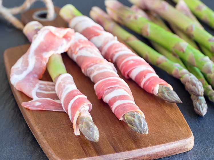 Raw asparagus spears getting wrapped in raw bacon slices for cooking in the oven. 